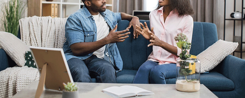 A couple sit on a couch while gesturing with their hands and arguing with one another. This could represent a relationship bump that a therapist in Ontario, Canada can help you address. Learn more about relationship fulfillment in Ontario, CA, and overcoming signs of an unfulfilled life.