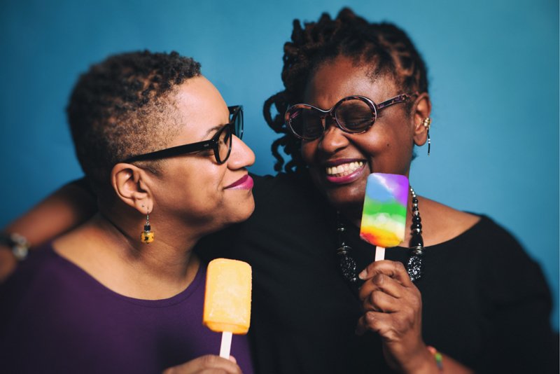 Two Black people standing close together in front of a blue background.  The person on the right is smiling and has twisted black hair that goes to their chin.  They are wearing round shape tortoiseshell sunglasses, and a black and gold earring.  They are wearing a black shirt and black shiny bead necklace.  In their left hand is a rainbow coloured popsicle.  They have their other arm around a second person to their right.  The second person is smiling and gazing back.  They have very short curly hair, is wearing black glasses with a yellow dangly earring.  They have on a purple shirt and a yellow popsicle in their hand.  