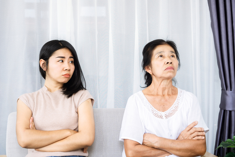 Two East Asian people stand apart looking tense in front of a white background.  Person on the right has an annoyed expression and is looking away with arms crossed at the chest.  They are wearing a white shirt, have dark shoulder length hair and looks to be in their sixties.   The second person next to them is looking back with a tense and frustrated expression, and arms crossed at chest.  They have black shoulder length hair, are wearing a beige shirt, and looks to be in their twenties.  