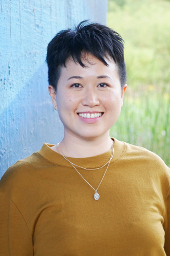 Jenny is an East Asian person with short black hair and wearing a mustard coloured sweater with two gold necklaces. They are smiling and standing in front of a blue wall with greenery to the side.