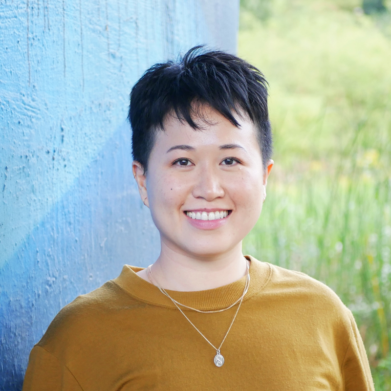 Jenny is an East Asian person with short black hair and wearing a mustard coloured sweater with two gold necklaces. They are smiling and standing in front of a blue wall with greenery to the side.